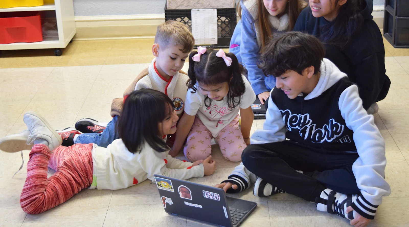 smiling students sit on the floor around a Chromebook while the older student presents